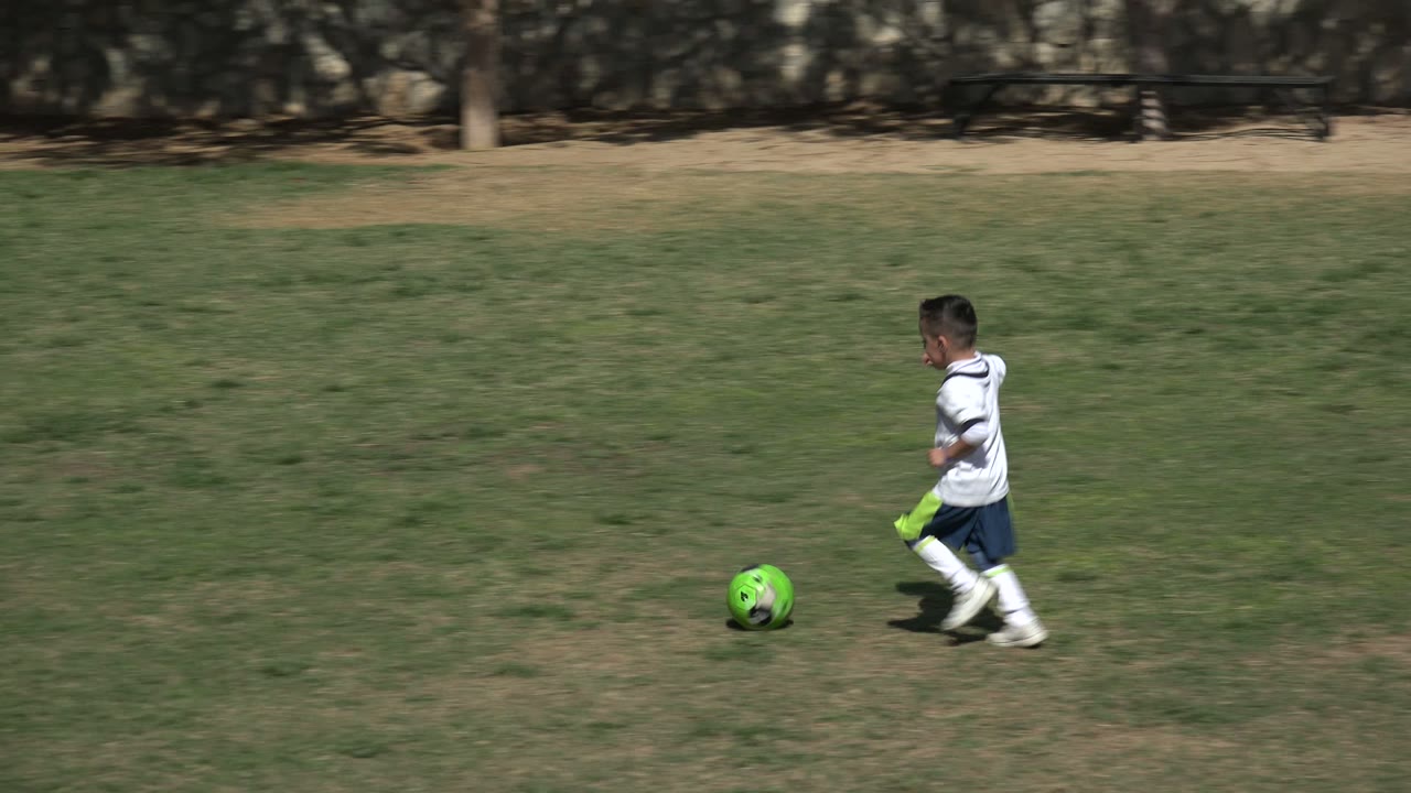 At 0:01, the boy moves toward the center of the grassy field in Los Cabos, Mexico, with the green soccer ball rolling just in front of him.