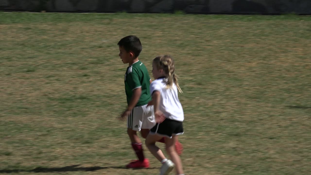 A child in a green jersey and a child in a white shirt run side by side across the Los Cabos soccer field under strong sunlight.