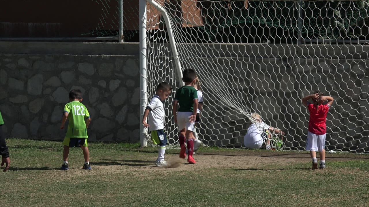 In Los Cabos, Mexico, children in green, white, and red soccer shirts stand around a white goal net while an adult coach approaches from the foreground.