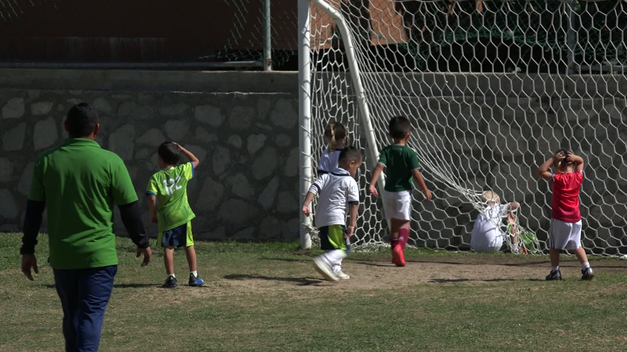 Children remain gathered at the soccer goal in Los Cabos, Mexico, with one child in red standing hands-on-head and the coach centered with his back to camera.