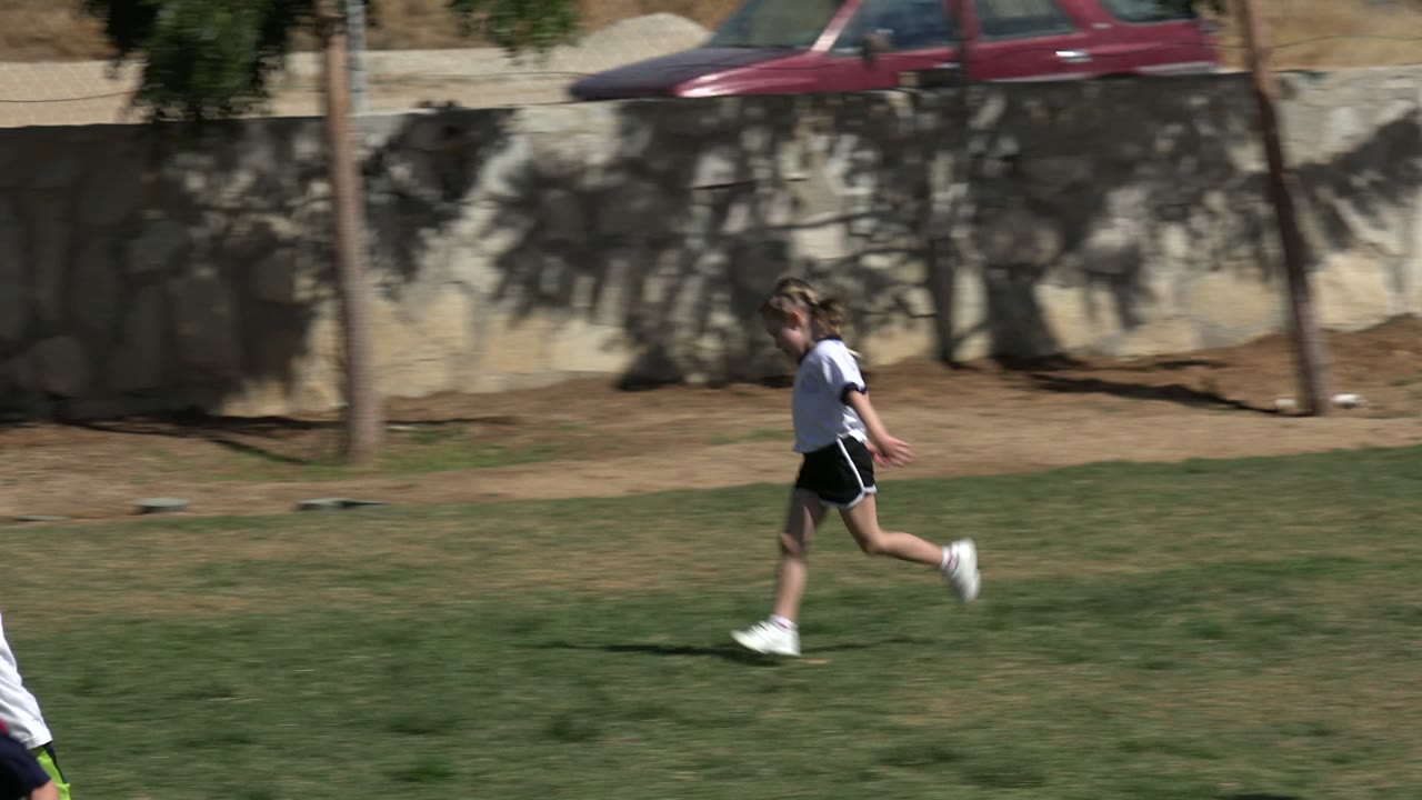 Young girl running across a grass soccer field near a stone wall.