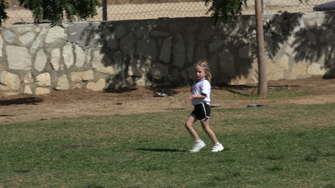 One second in, the girl is farther across the Los Cabos soccer field, seen in profile with patchy grass, tree shade, fencing, and a stone wall in the background.