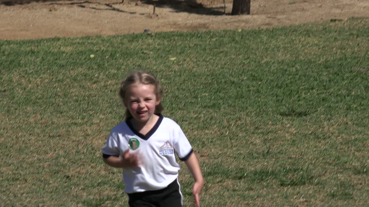 A girl in a white soccer-style shirt runs on the grass in Los Cabos, Mexico, with a boy's head partly visible in the foreground and dry park ground behind her.