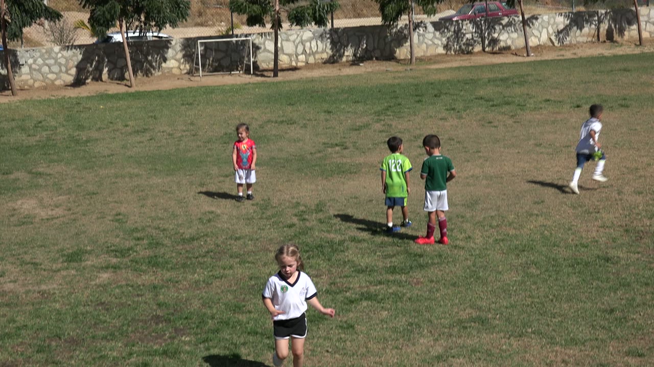Midwide frame of children in green and white sports clothing standing on the Los Cabos football field, with one child in red farther back and goals near the wall.