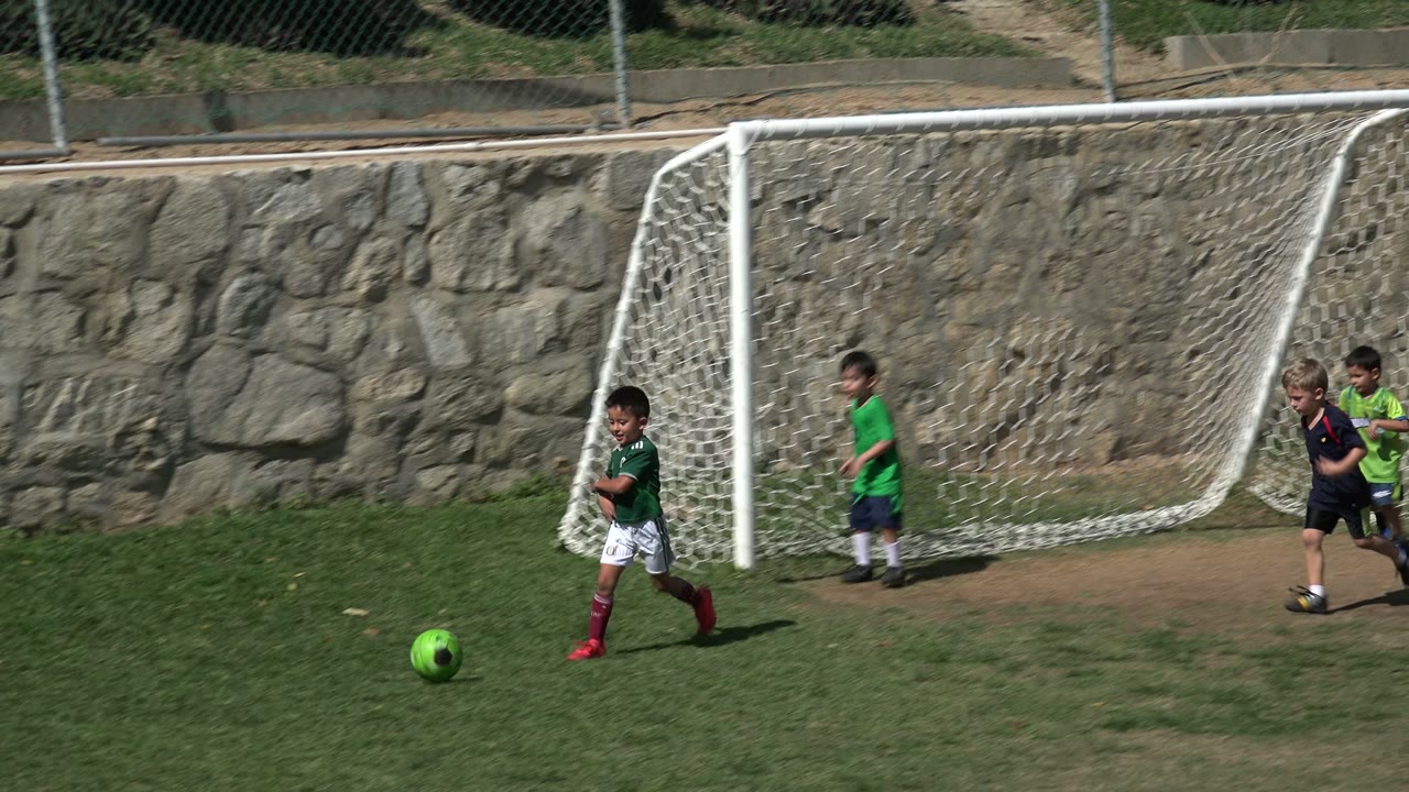 A child in a green jersey moves toward the bright green soccer ball on the Los Cabos field while two other children run near the white goal net.
