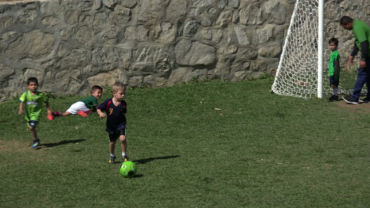 At 0:01 in Los Cabos, Mexico, a blond child in a dark uniform dribbles the green soccer ball while other children and the goal net remain in the background.
