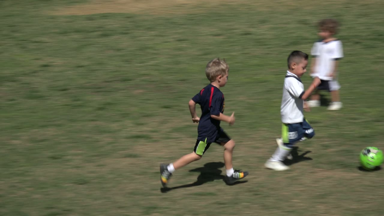 Three boys in sports uniforms chase a bright green football across a sunlit grass field in Los Cabos, Mexico, with the ball ahead of them.