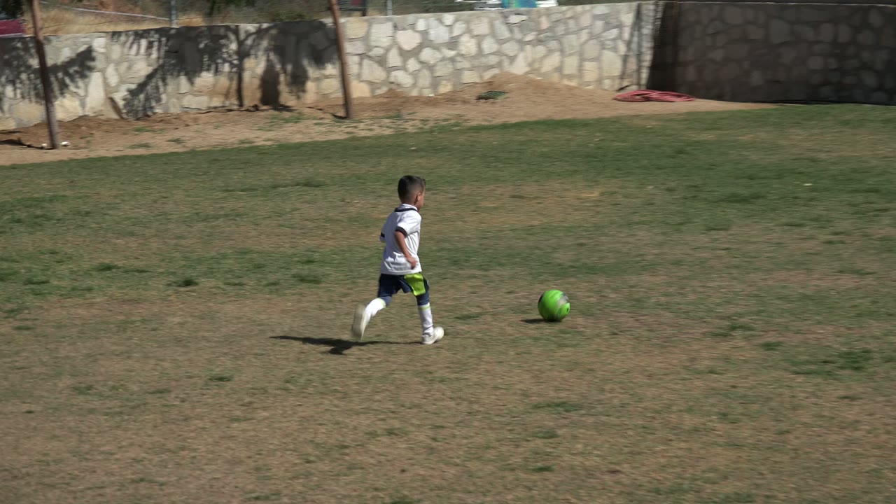 Young child in a white soccer uniform running toward a green ball on grass.
