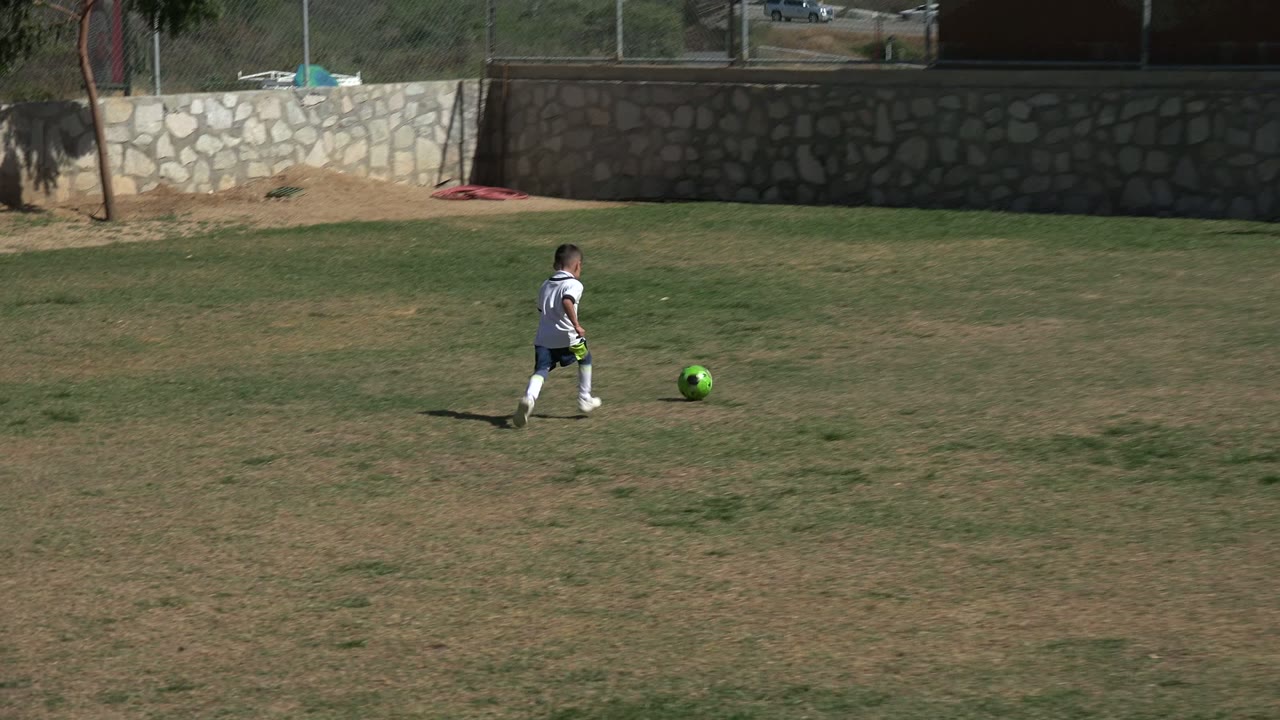 At 0:01, the child in white soccer gear moves farther across the Los Cabos grass field with the green ball near their feet and the stone wall still filling the background.