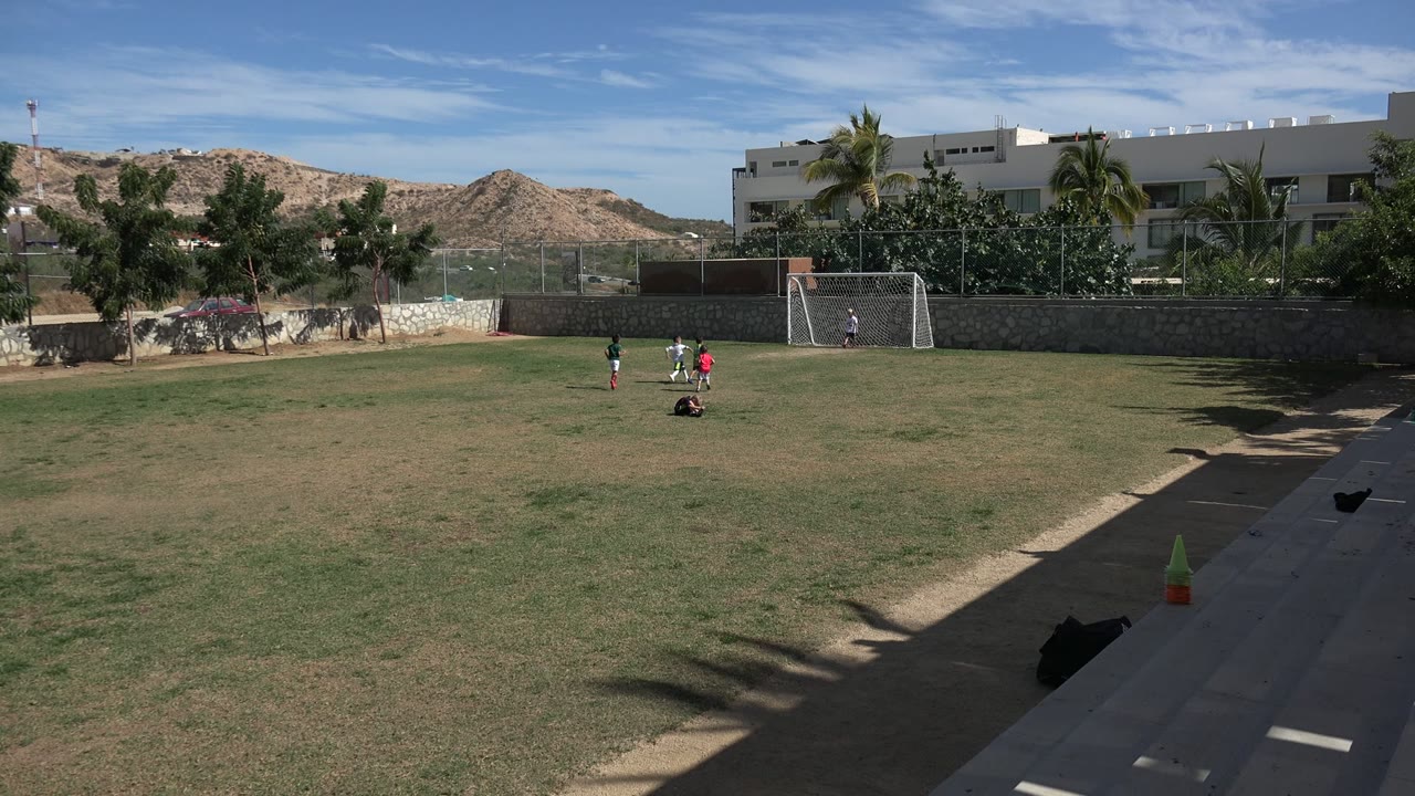 Wide soccer field with children near a goal and hills behind low buildings.