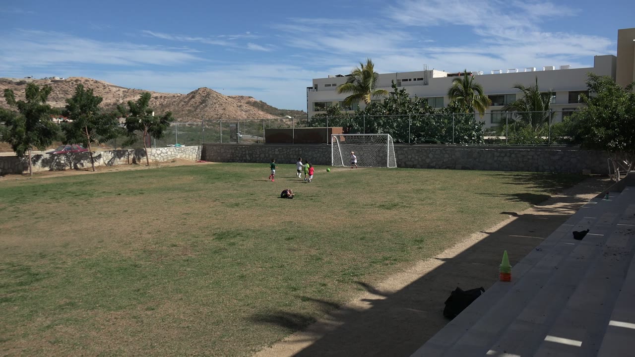 A wide view shows children moving near the goal on the Los Cabos soccer field, with one person low on the grass and a shaded walkway in the foreground.