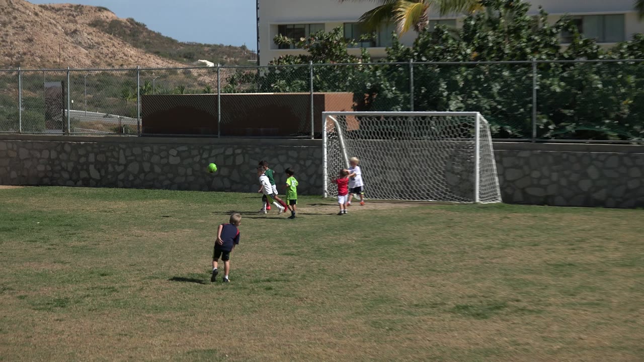Children play football near a metal goal on a patchy grass field in Los Cabos, Mexico, with a stone wall, fence, road, and hillside behind them.