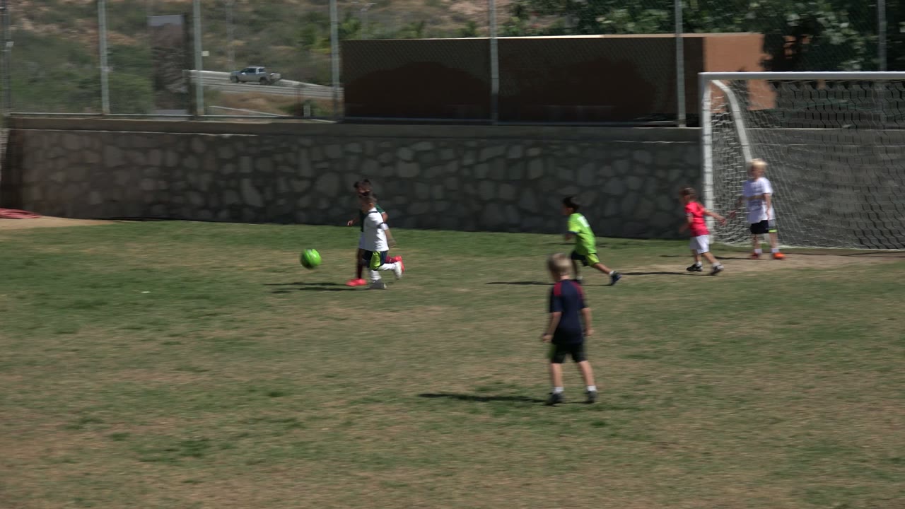 Children in sportswear chase a bright green football across the Los Cabos grass field, with the goal now farther left and the stone wall in the background.