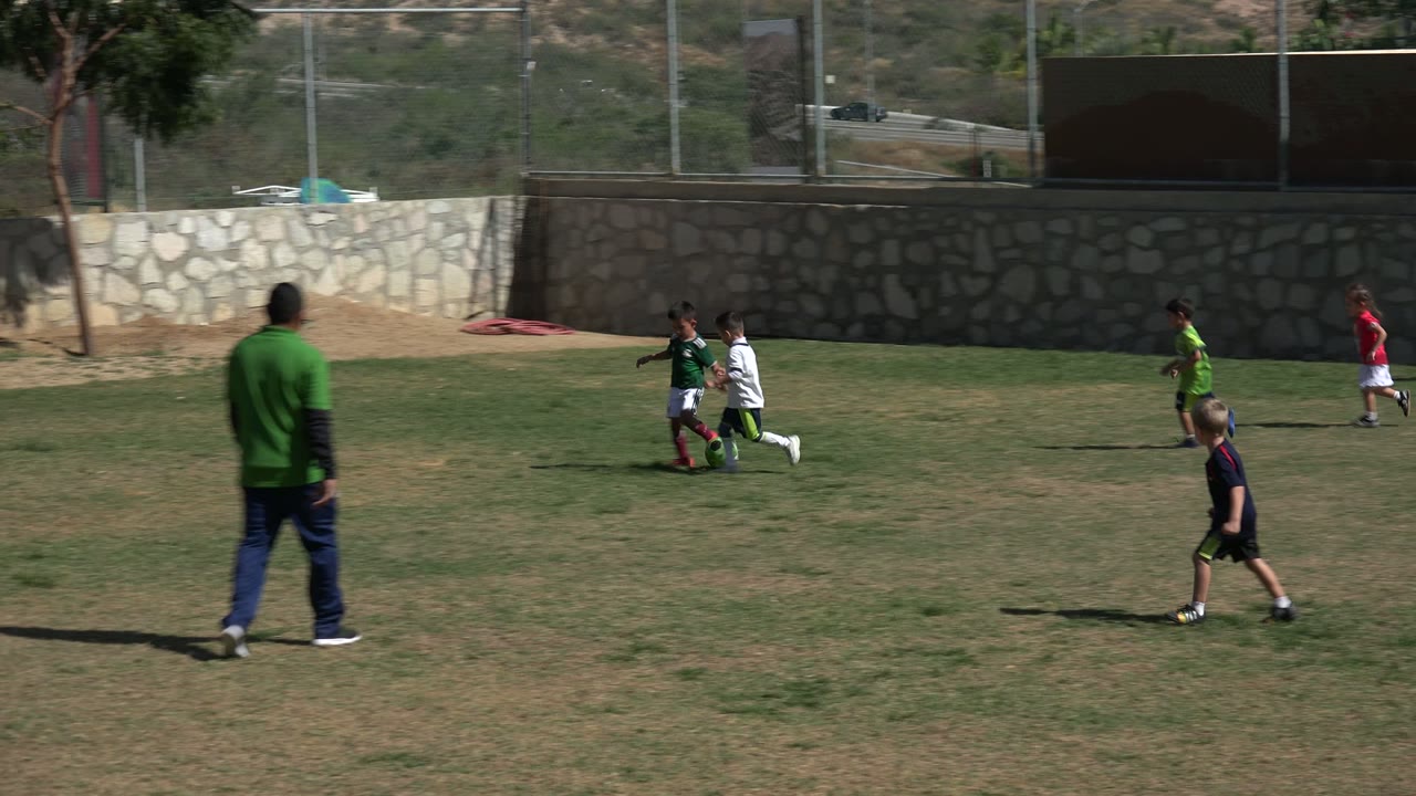 An adult stands on the field while children play football nearby.