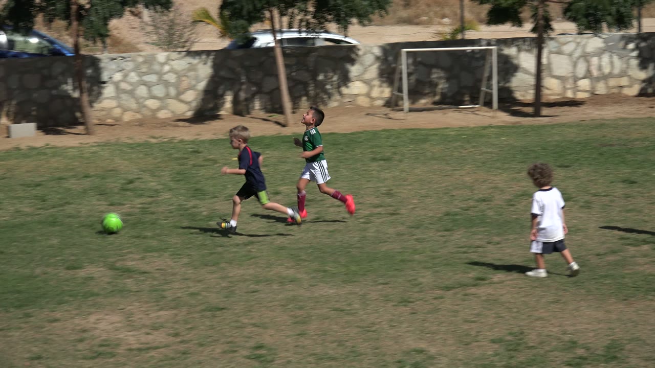 Boys run across the sunlit soccer field in Los Cabos, Mexico, with one green ball near the center and the stone wall and cars in the background.