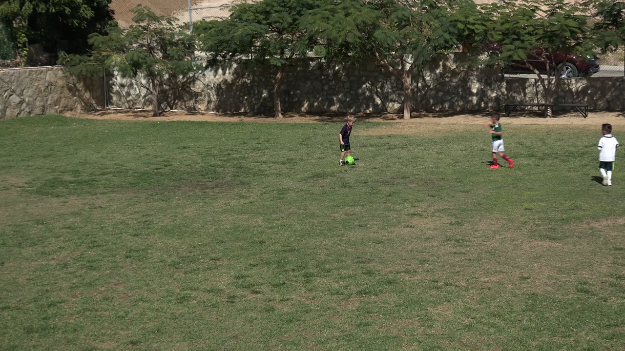 In Los Cabos, Mexico, two children in soccer uniforms stand apart on a patchy grass field with trees, a stone wall, and parked vehicles behind them.