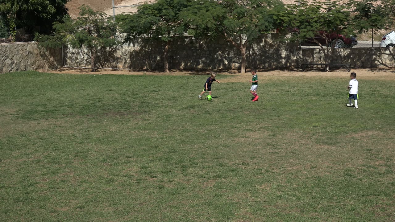 In Los Cabos, Mexico, several children are spaced across the field while one child in a dark uniform approaches the bright green soccer ball near the center.