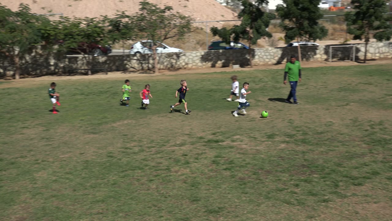 Children playing soccer on a grassy field with an adult nearby.