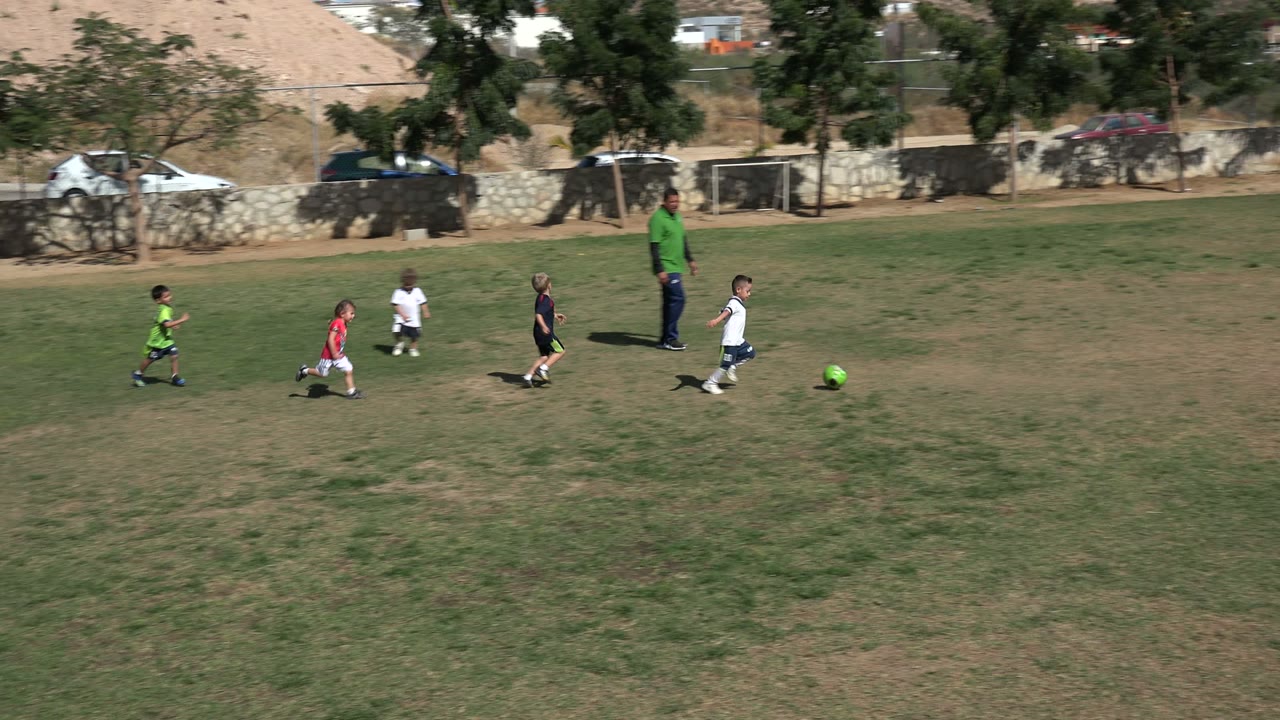Children in red, white, black, and green soccer outfits run after a bright green ball on the Los Cabos field, with a coach standing farther back near the wall.