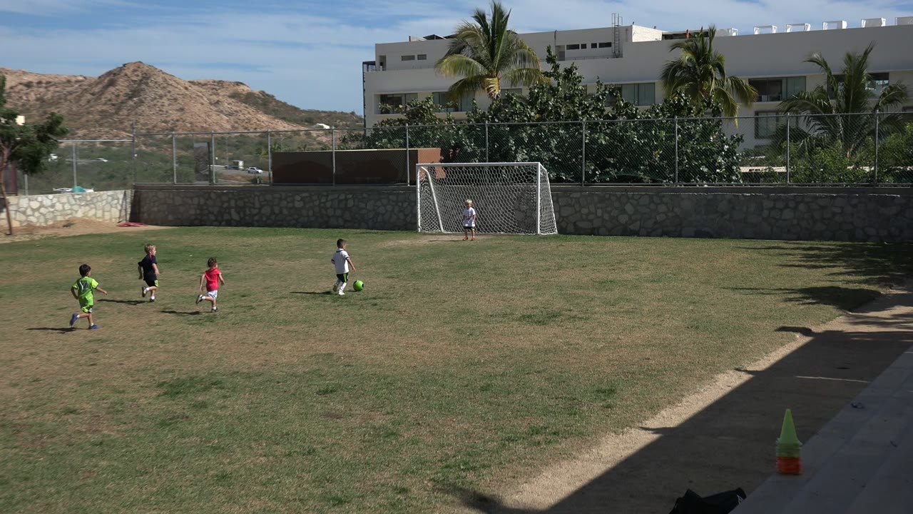 Wide view in Los Cabos, Mexico, with children spread across a sunlit grass field as a green soccer ball sits near a child in white and a white goal stands behind them.