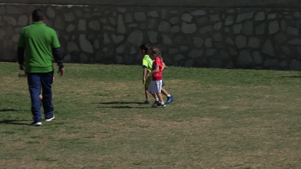 In Los Cabos, Mexico, an adult in a green shirt stands at the left while several children gather on a grass soccer field near a stone wall and fence.