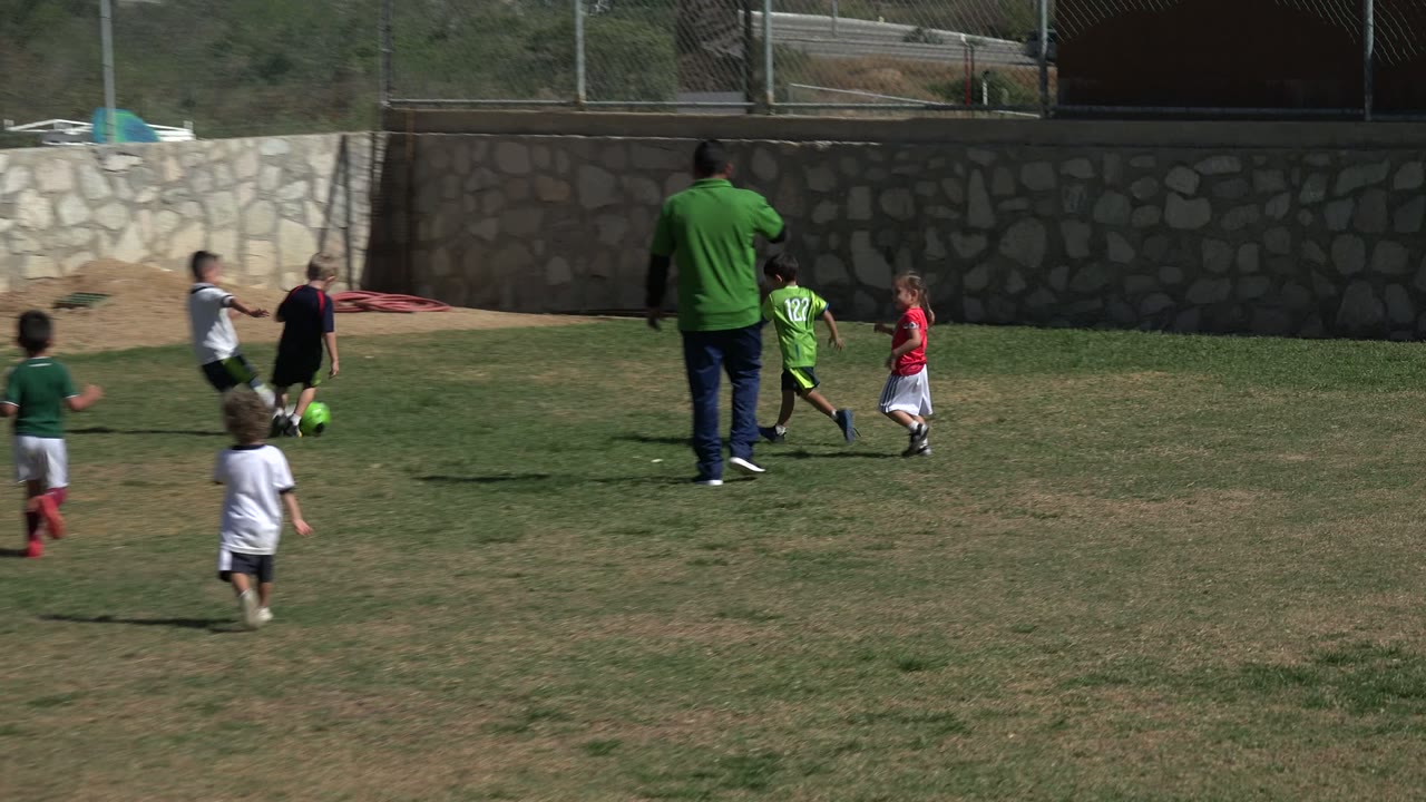 In Los Cabos, Mexico, children run toward a bright green soccer ball as the adult coach stands in the midground beside the play.