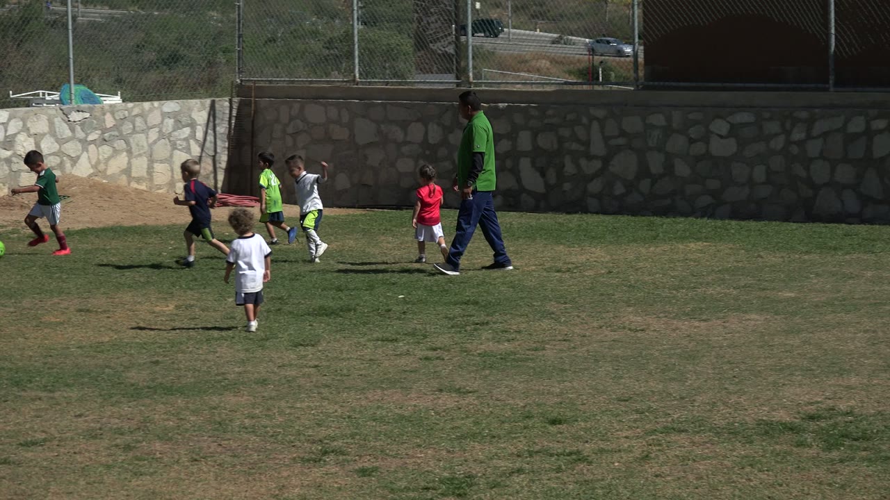Several children run after the ball in Los Cabos, Mexico, with one child in a red shirt near center and the adult in green walking beside the group.