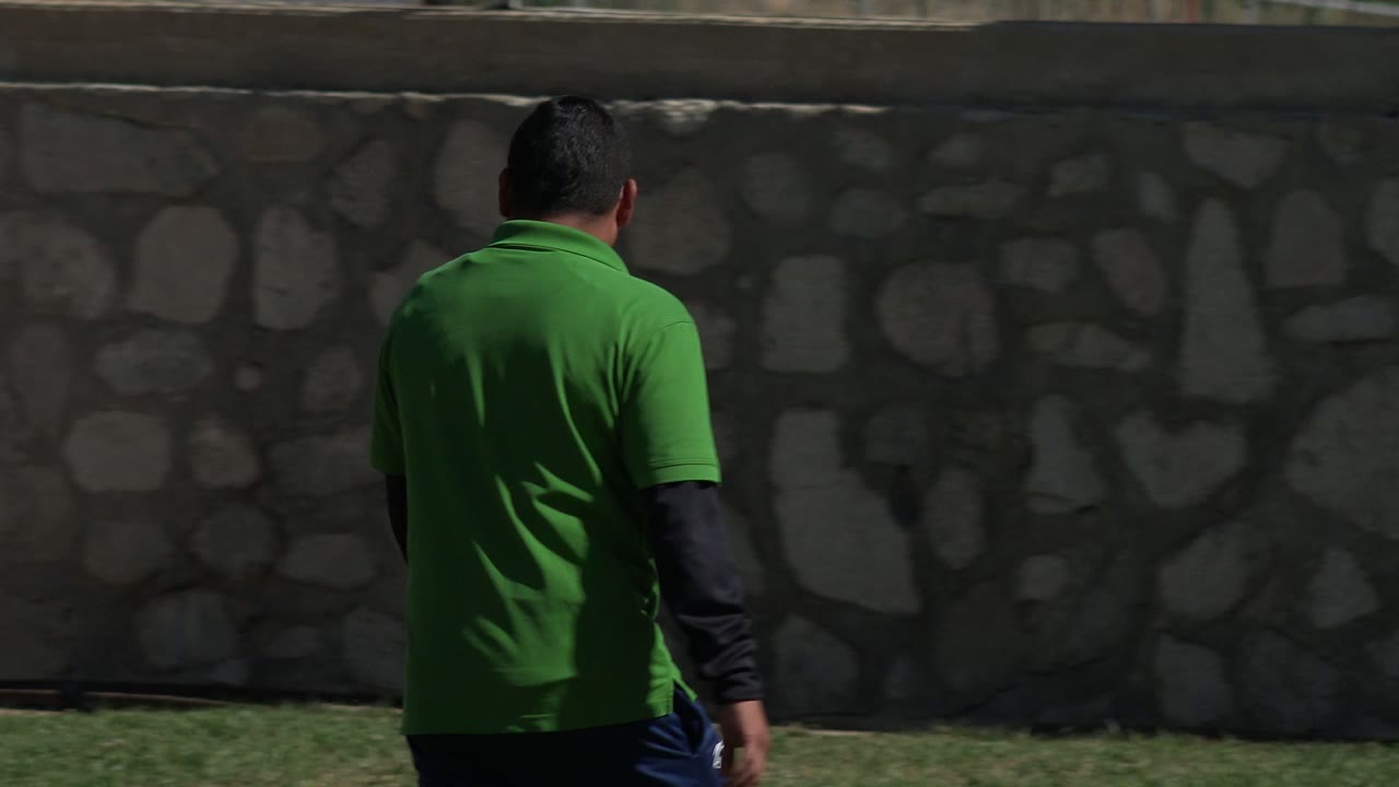 In Los Cabos, Mexico, the man in the bright green shirt appears more centered with his back to camera, walking parallel to the long stone barrier.