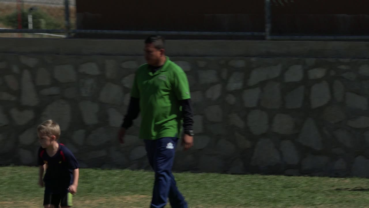 In Los Cabos, Mexico, a young boy in a dark shirt runs across the grass while an adult coach in a green polo stands nearby against a stone wall and fence.