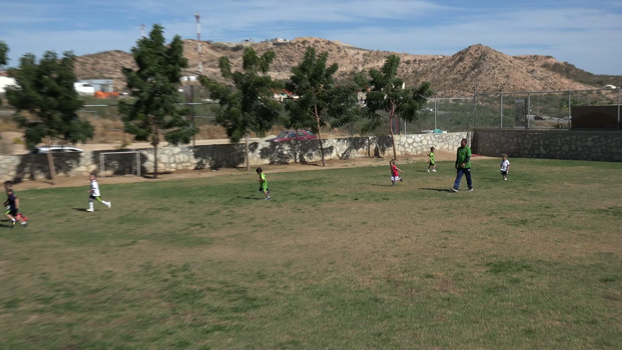 One second in, children move across the Los Cabos grass field around a small ball while an adult in a green shirt stands near the center and dry hills line the background.