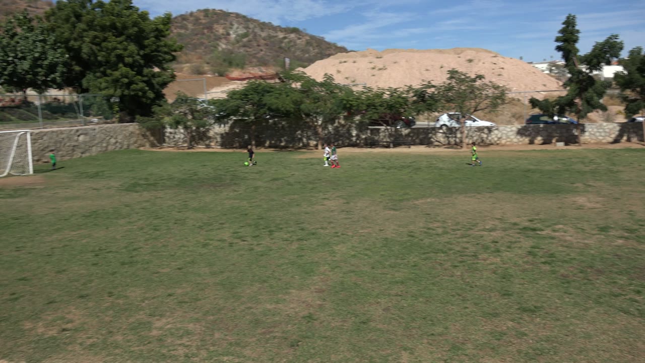 Wide soccer field with distant players, goal, trees, and hills.