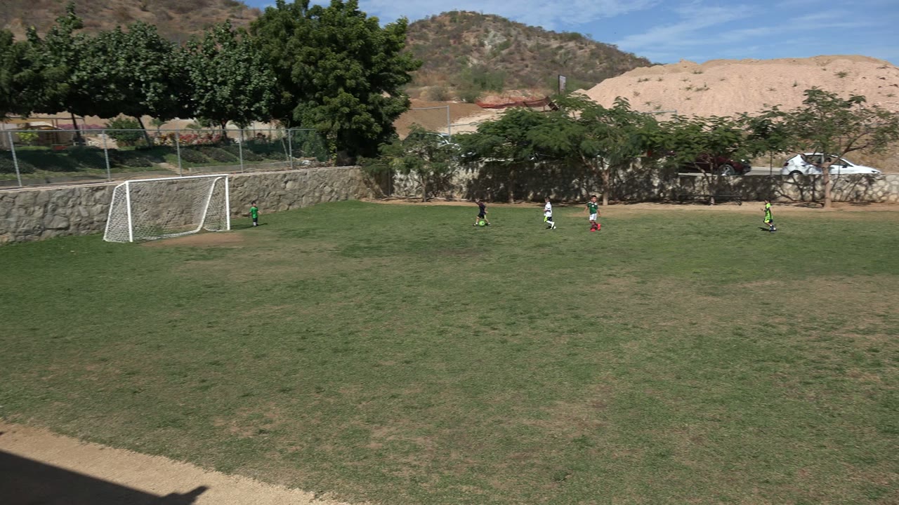 A Los Cabos soccer field remains in a wide view as small players stand and run near the white goal, with trees and tan earth banks behind the wall.