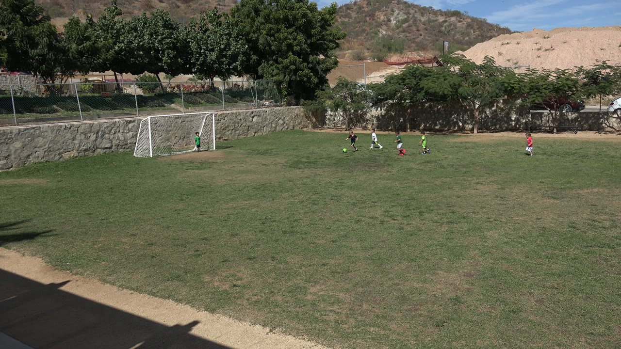 Children play soccer near a white goal on a grassy field in Los Cabos, Mexico, with trees, fencing, and low hills behind them.