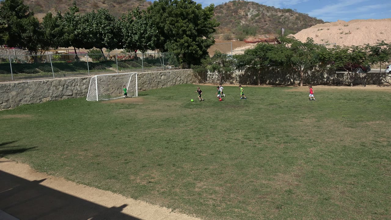 A small group of children runs after a bright green soccer ball near the left-side goal in Los Cabos, Mexico, while another child stands farther right.