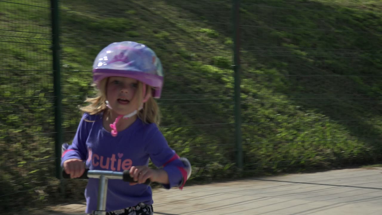 The girl rides closer toward the camera in Los Cabos, holding the scooter handlebars with the shaded fence line and path behind her.