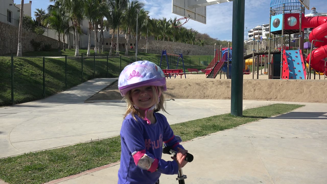 Young girl in a purple helmet smiling beside a scooter in a park.