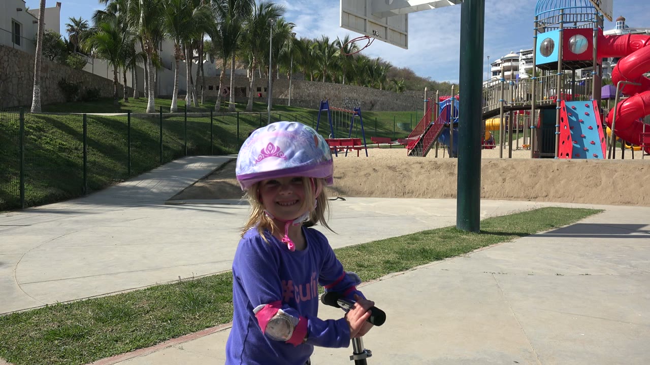 One second in, the helmeted girl remains in the foreground of the Los Cabos park, smiling toward the camera with red playground slides and palms in view.
