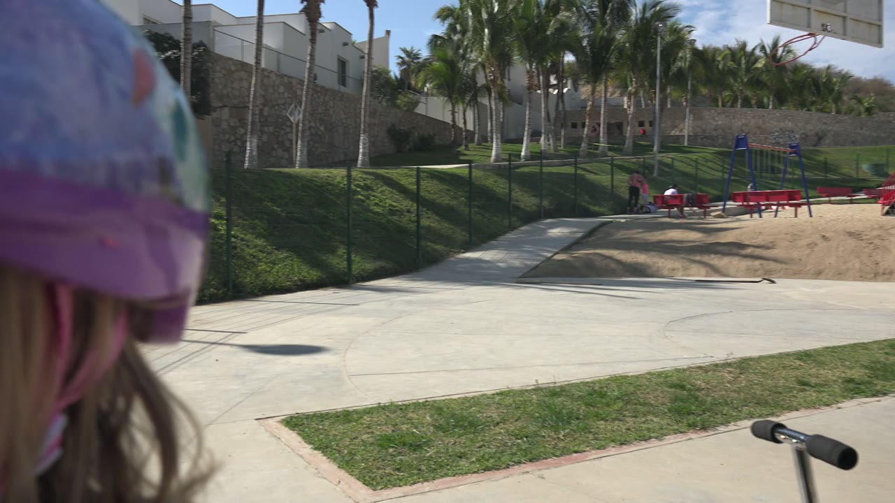 At the start of the Los Cabos clip, part of a young girl in a purple helmet fills the left foreground beside a concrete park path, grass, fencing, palms, benches, and buildings.