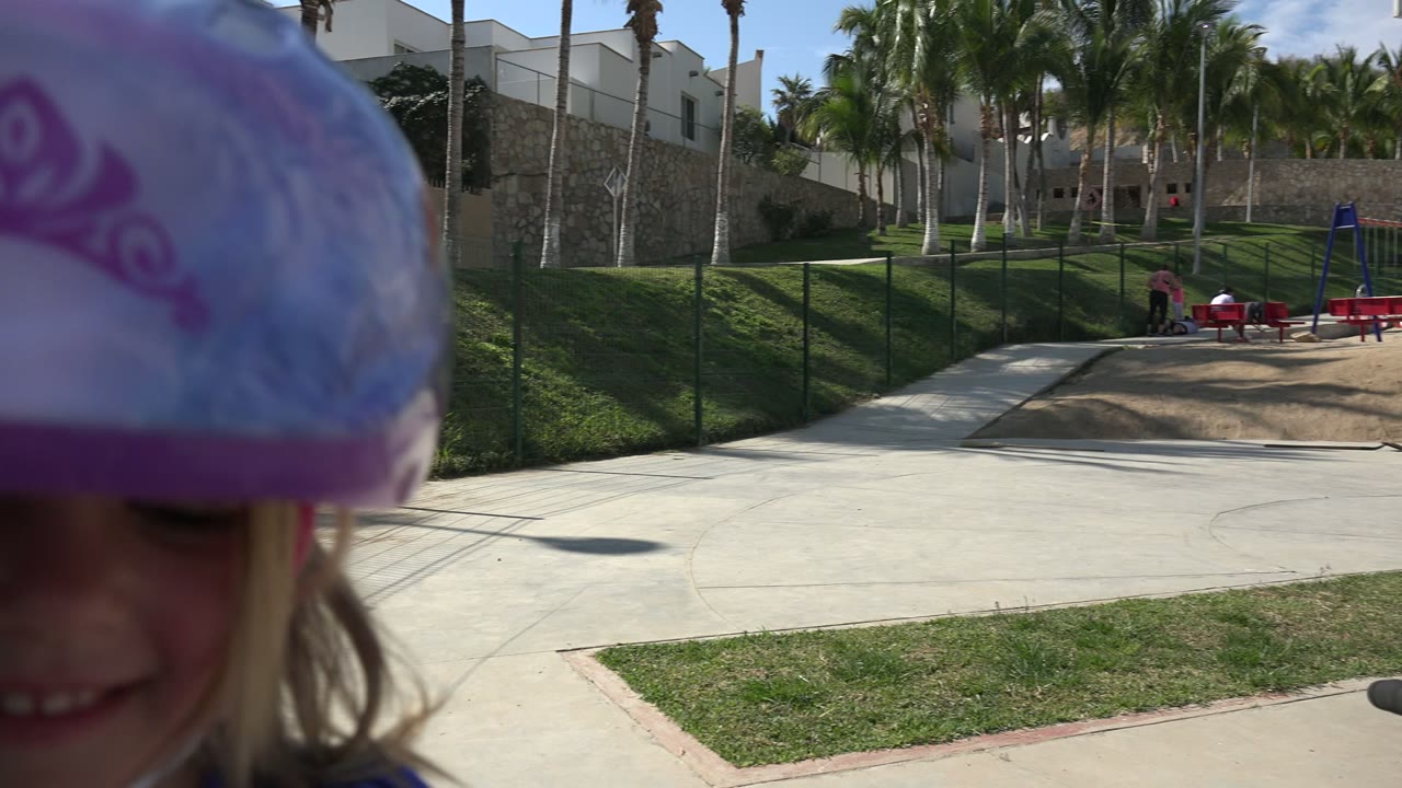 A young girl in a purple helmet smiles close to the camera in Los Cabos, with a scooter handle in front of her and a fenced park path, grass, benches, palms, and buildings behind.