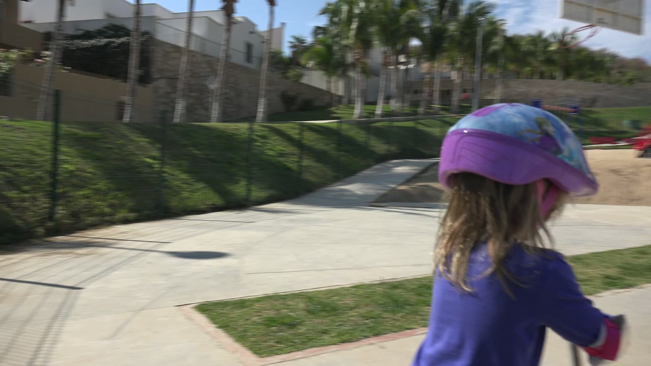 A child in a purple helmet rides a scooter along a paved area in Los Cabos, Mexico, with grass, fencing, and playground equipment ahead.