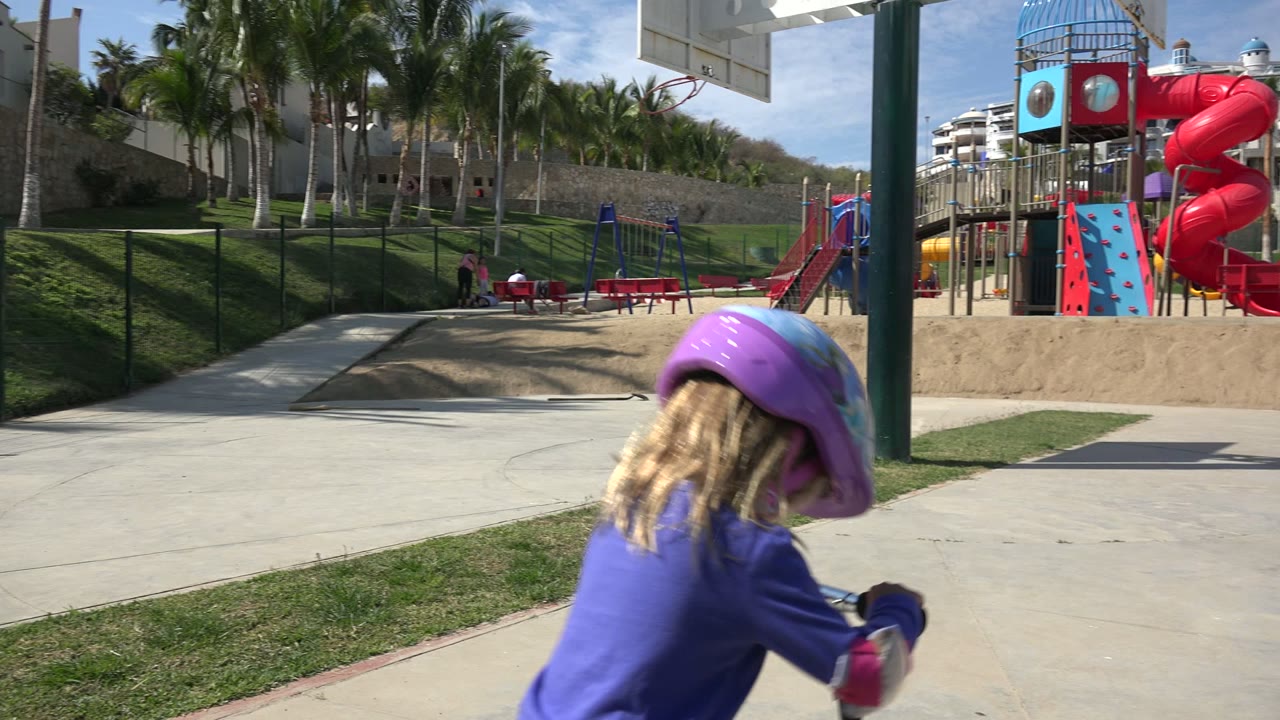 The child continues forward on a scooter in Los Cabos, Mexico, with a red spiral slide, climbing structure, swings, palms, and hillside homes in the background.