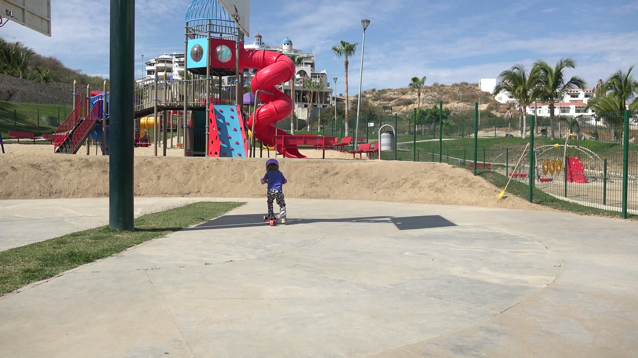 Child riding a scooter on a park path near a playground.