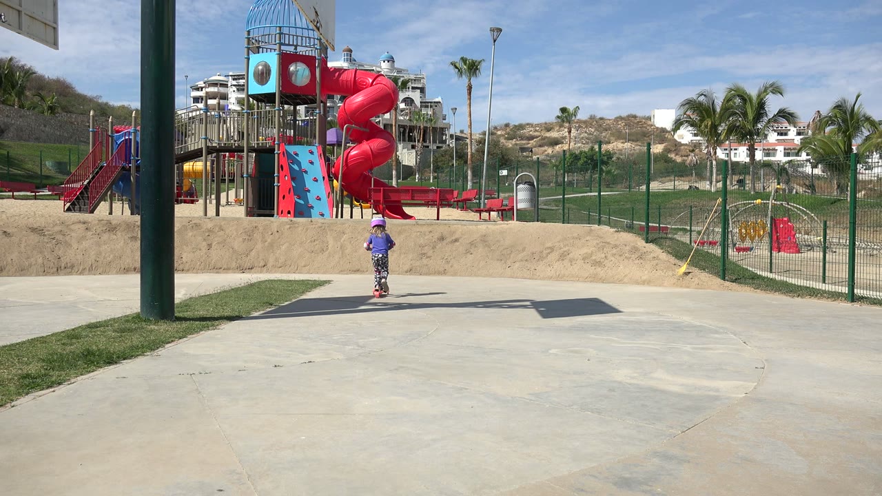 The child continues riding a scooter near the center of the paved path in Los Cabos, framed by a tall green pole, sand berm, red slide, and palms.