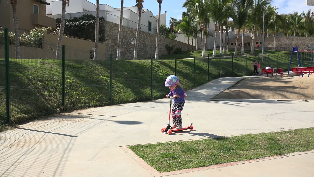 A young helmeted girl rides a red scooter across a paved path in Los Cabos, with grass, fencing, palm trees, and hillside buildings behind her.