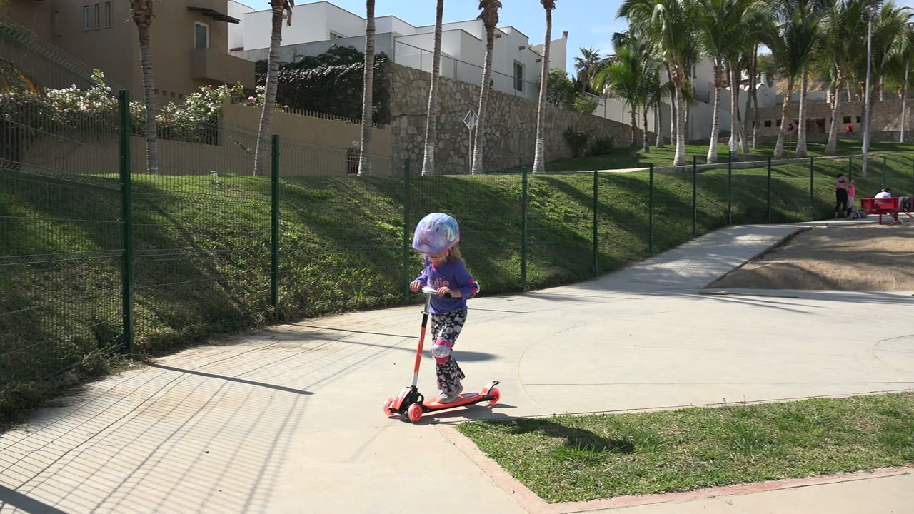 The child continues riding the red scooter in Los Cabos, positioned near the grass edge with the fenced walkway and palms filling the background.
