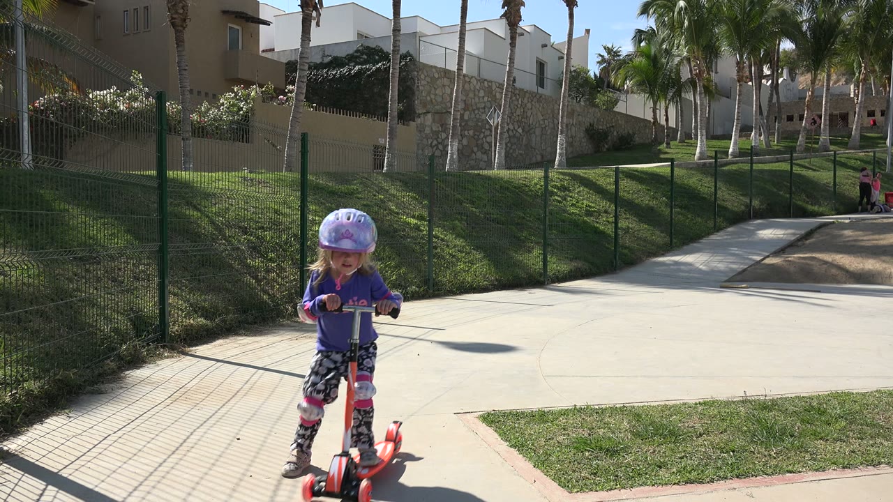 Girl in purple shirt and helmet rides toward the camera on a red scooter.