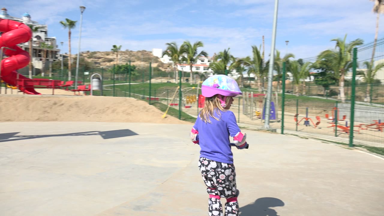 Young girl in helmet standing with scooter near a fenced playground
