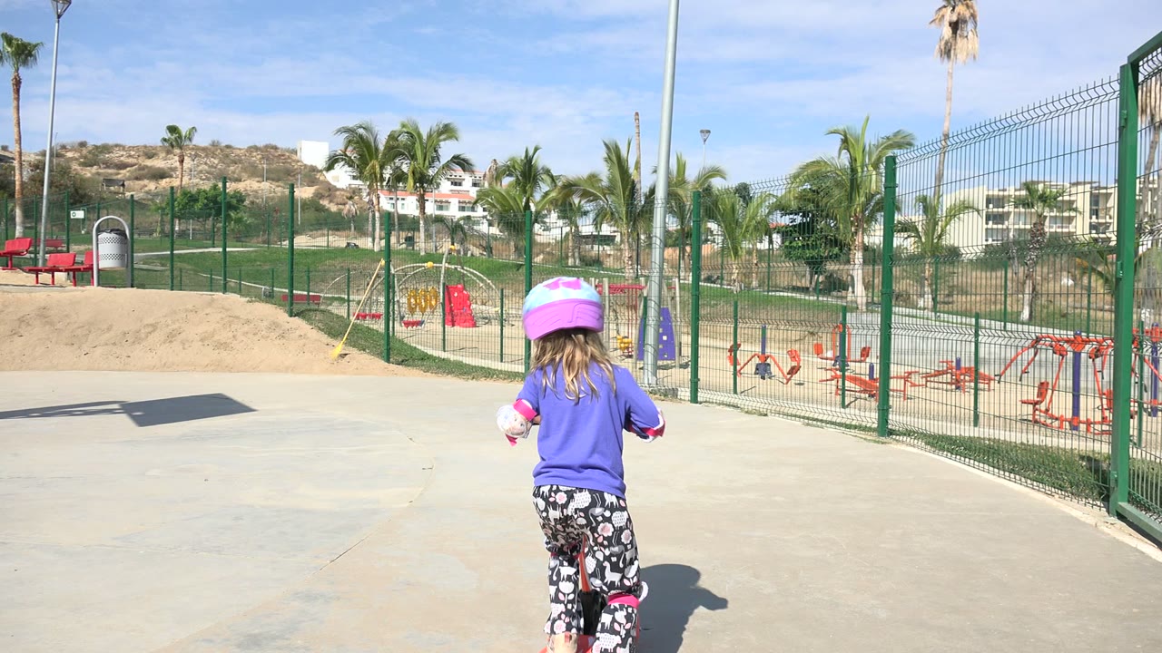 The girl is seen from behind as she starts forward on the orange scooter across the paved playground, with green fencing and palms behind her in Los Cabos, Mexico.