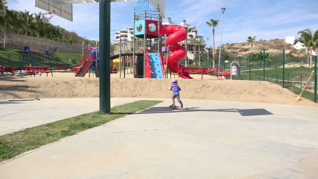 In Los Cabos, Mexico, a child in a purple shirt and helmet rides an orange scooter along a concrete sidewalk in front of a red playground slide and sandy play area.