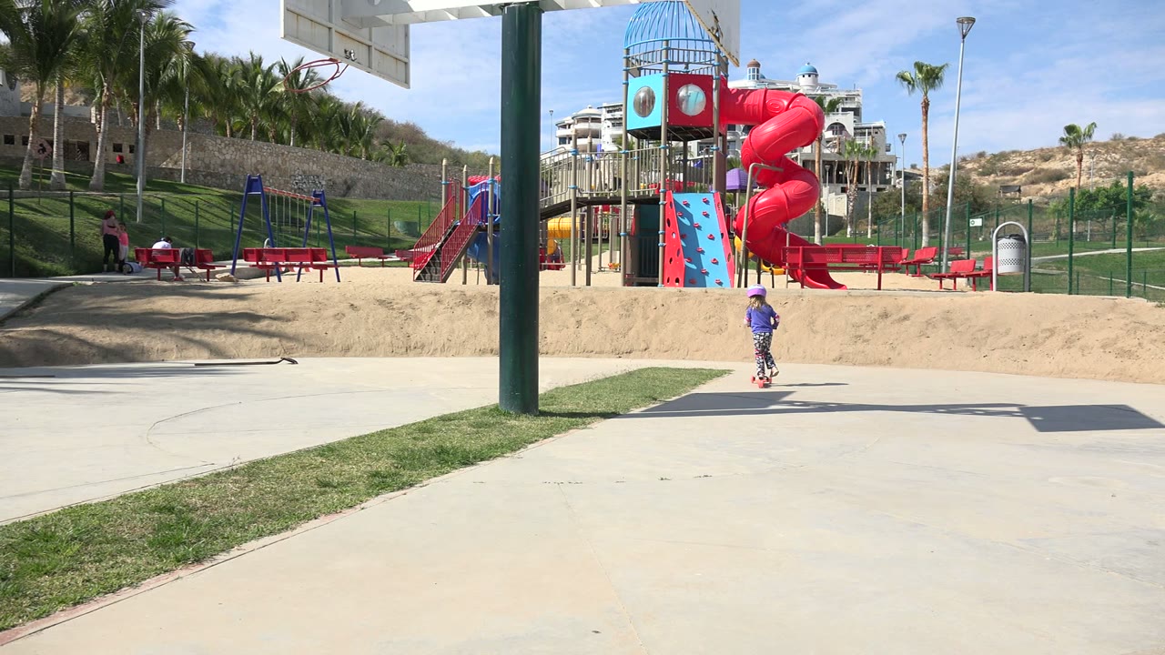 A wide Los Cabos playground view shows swings, a red slide, benches, palm trees, hillside buildings, and small visible people near the shaded play area.